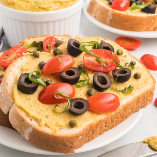 two slices of bread with homemade garlic hummus without tahini, tomatoes, olives, fresh herbs, and capers.
