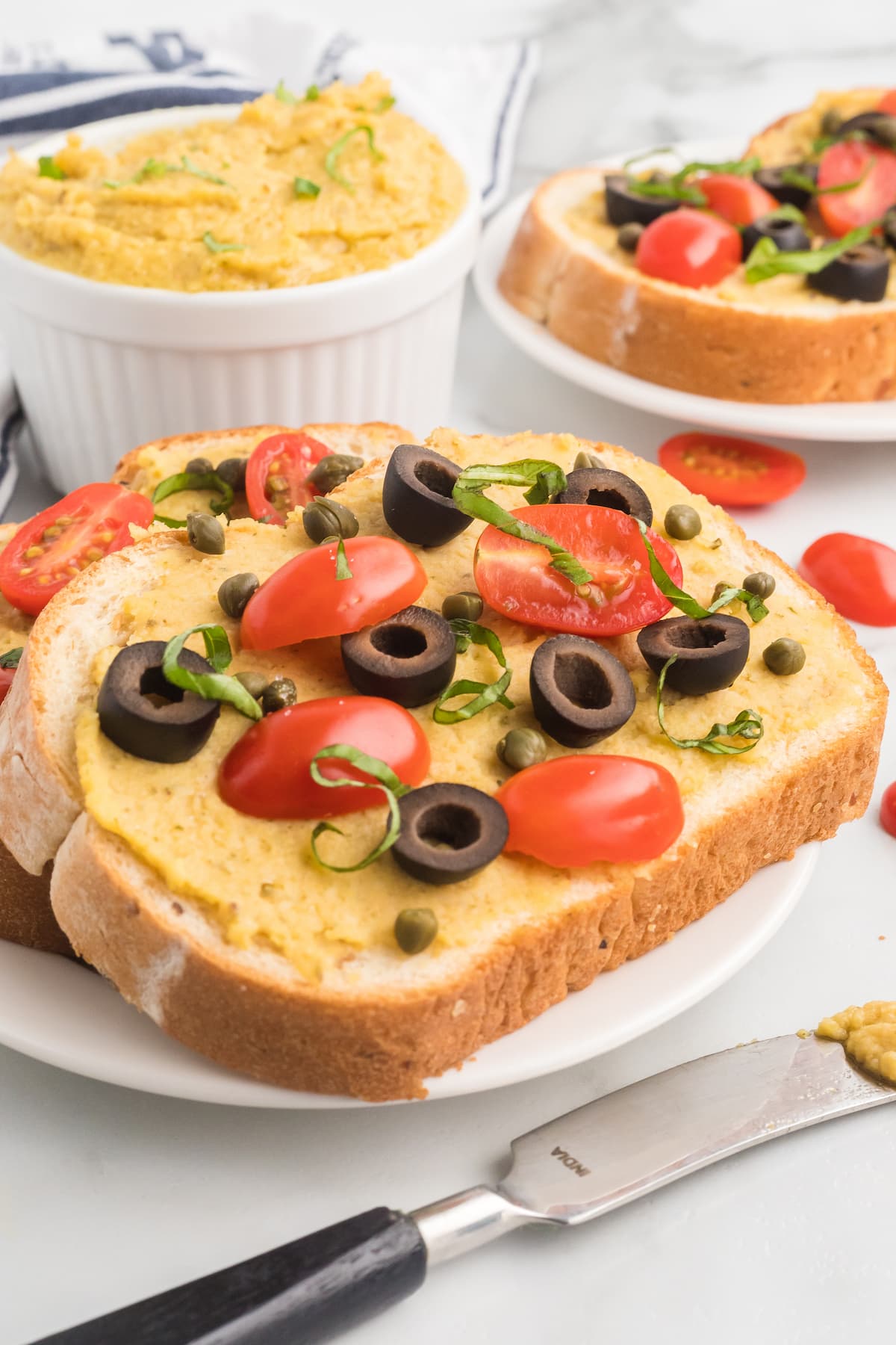two slices of bread with homemade garlic hummus without tahini, tomatoes, olives, fresh herbs, and capers. 