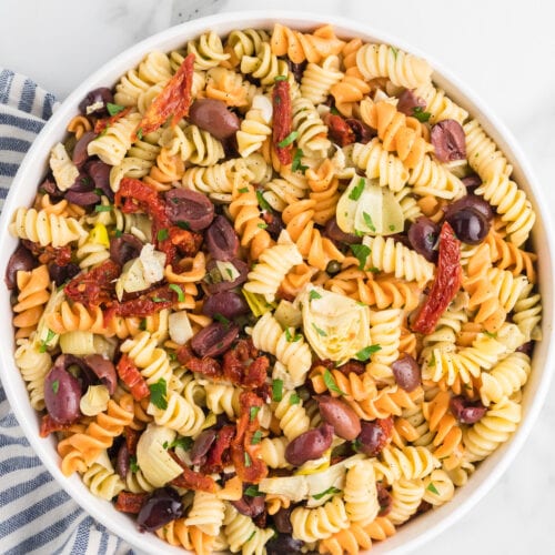 white serving bowl with the finished Mediterranean pasta salad and striped tea towel to the side.