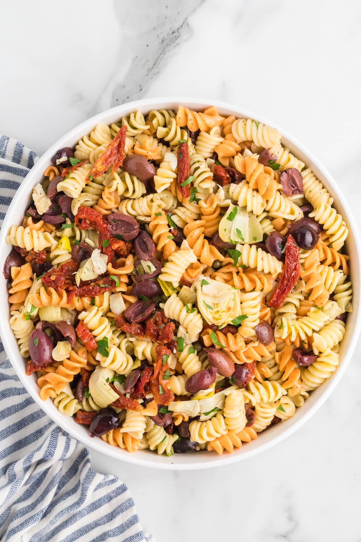 white serving bowl with the finished Mediterranean pasta salad and striped tea towel to the side.