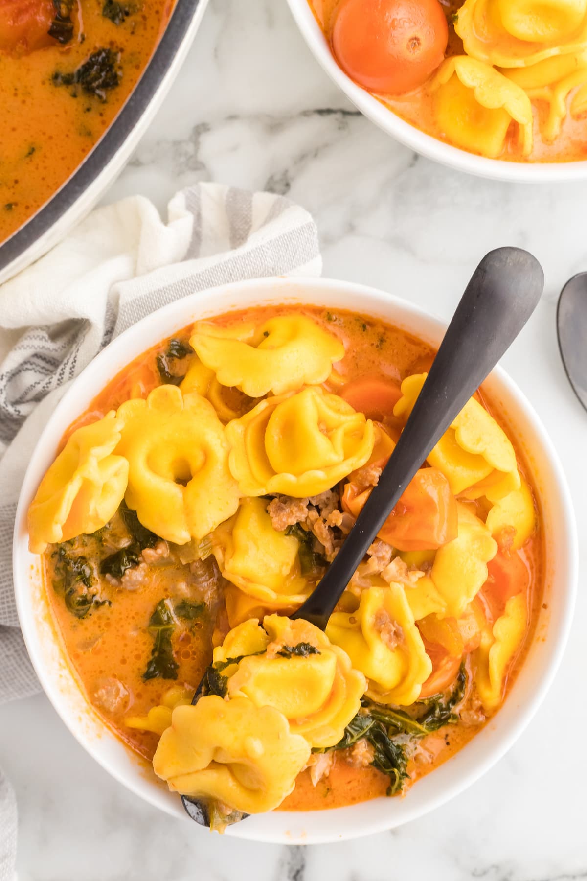 white bowl of creamy italian sausage soup with tortellini and a black spoon inside of the bowl.