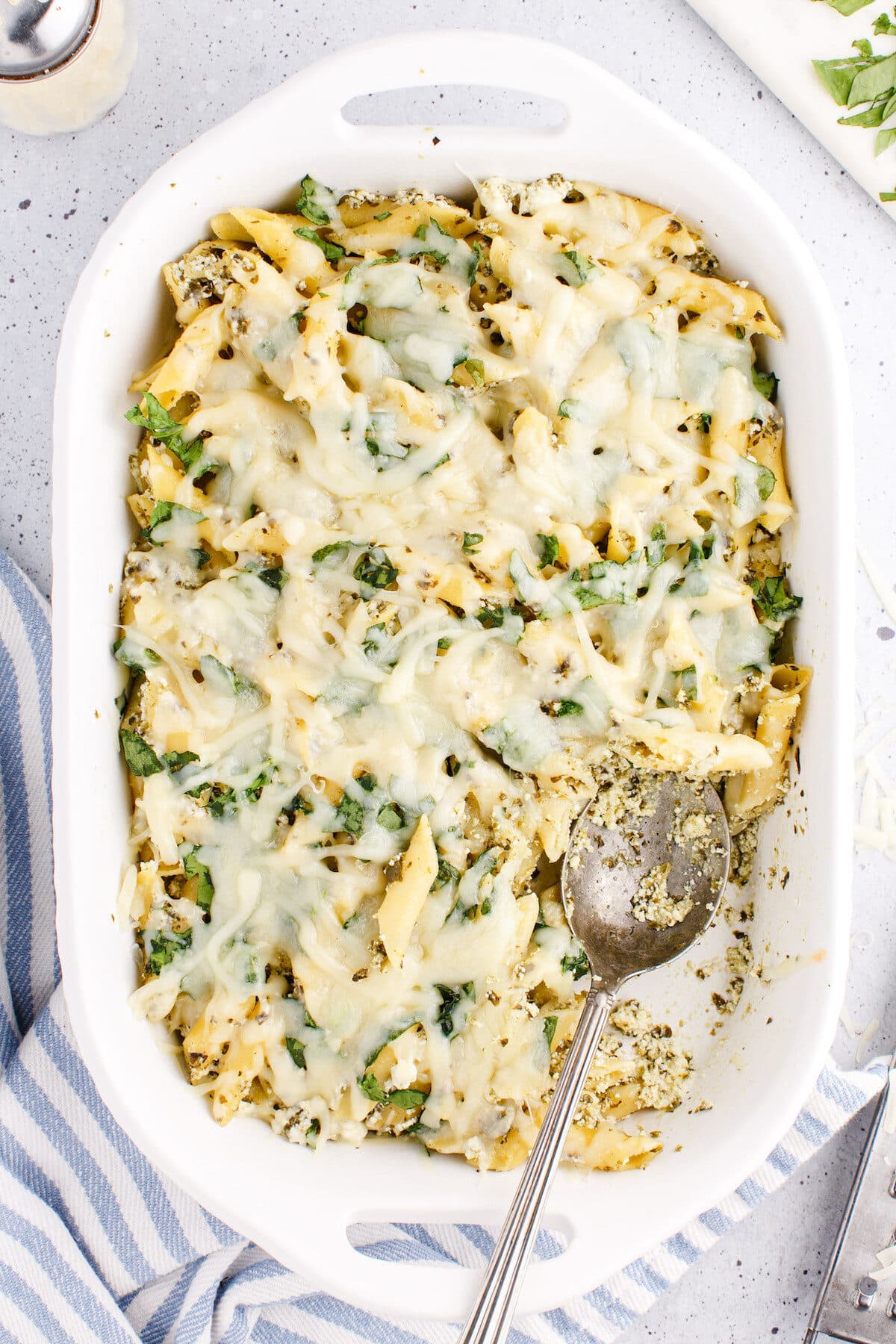 white casserole dish with baked pesto pasta and a serving spoon to the side.