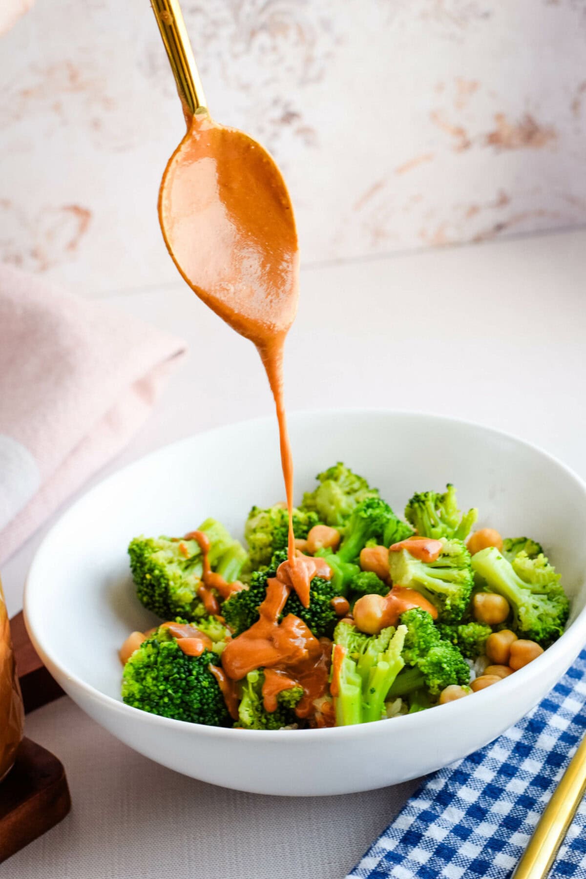 drizzling the Thai peanut sauce over a white bowl filled with brown rice, broccoli, chickpeas. 